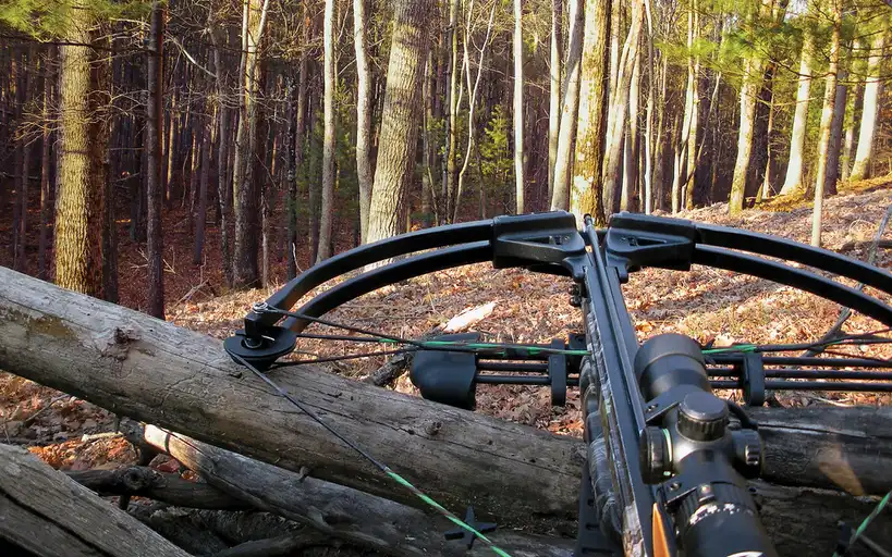 Gloved hands cocking a crossbow beside bolts and a broadhead in natural light